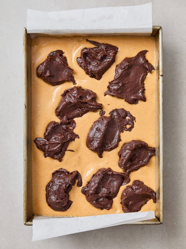 Dollops of chocolate brownie batter sit on top of a pumpkin cheesecake layer in a pan.