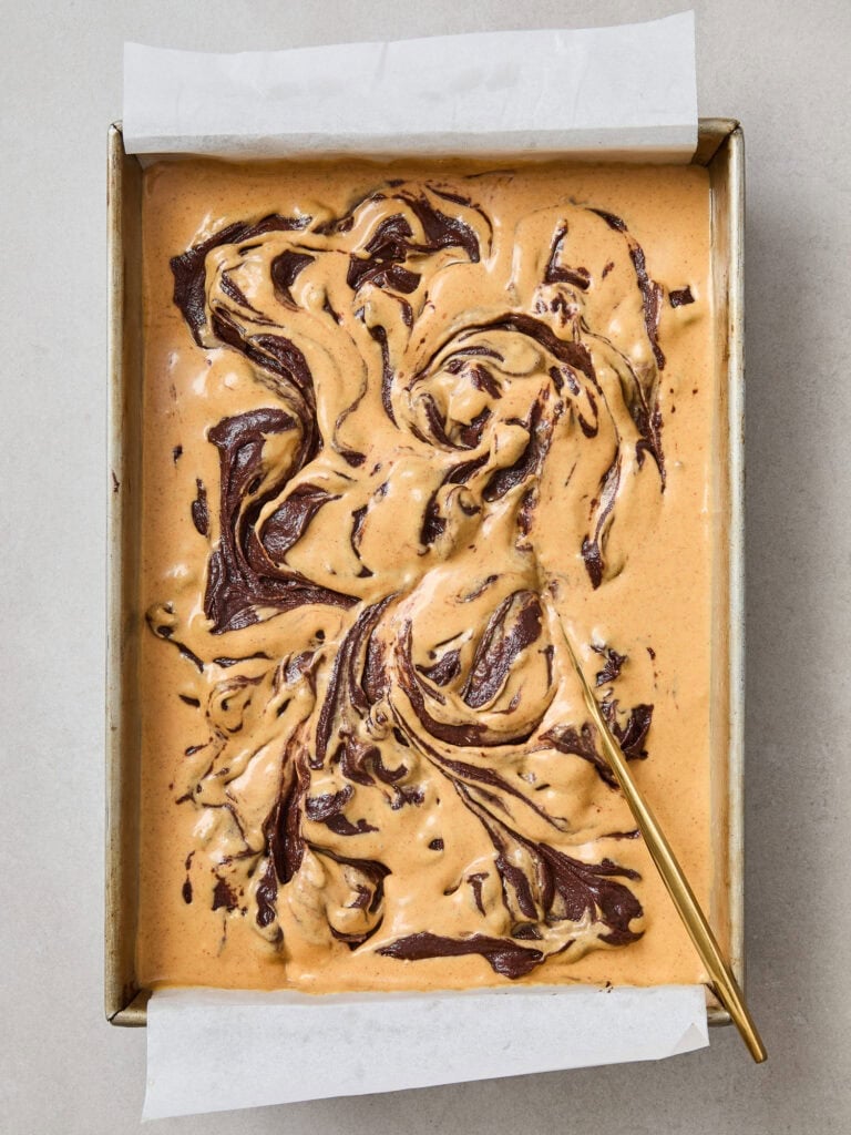 Pumpkin cheesecake and brownie batter in a pan after being swirled together.