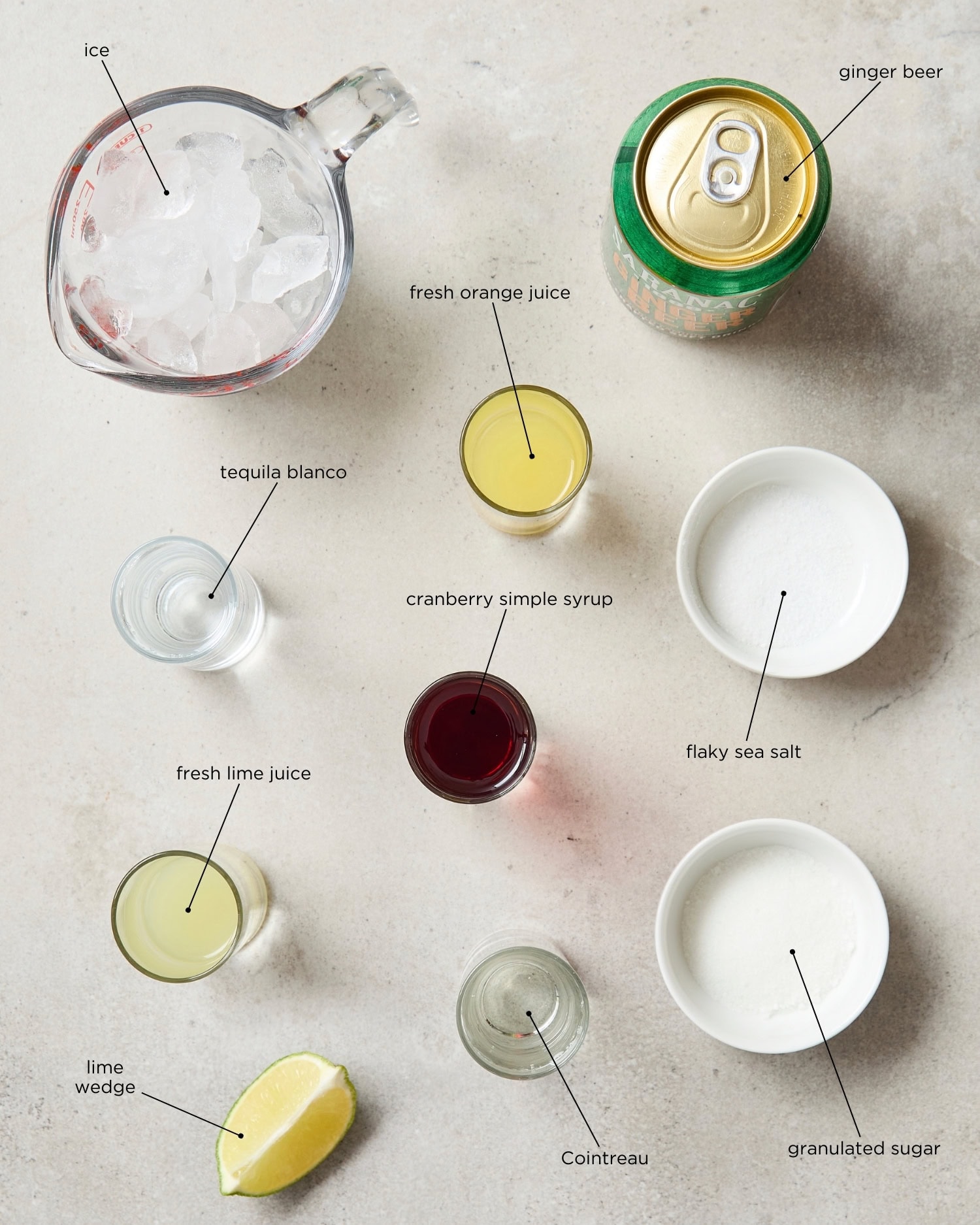 Concrete surface with ingredients for a cranberry margarita including ice, ginger beer, salt, sugar, and more.