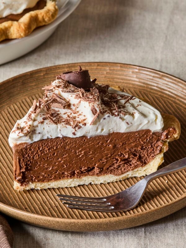 Slice of French silk pie with whipped cream and chocolate curls on top on a brown plate.
