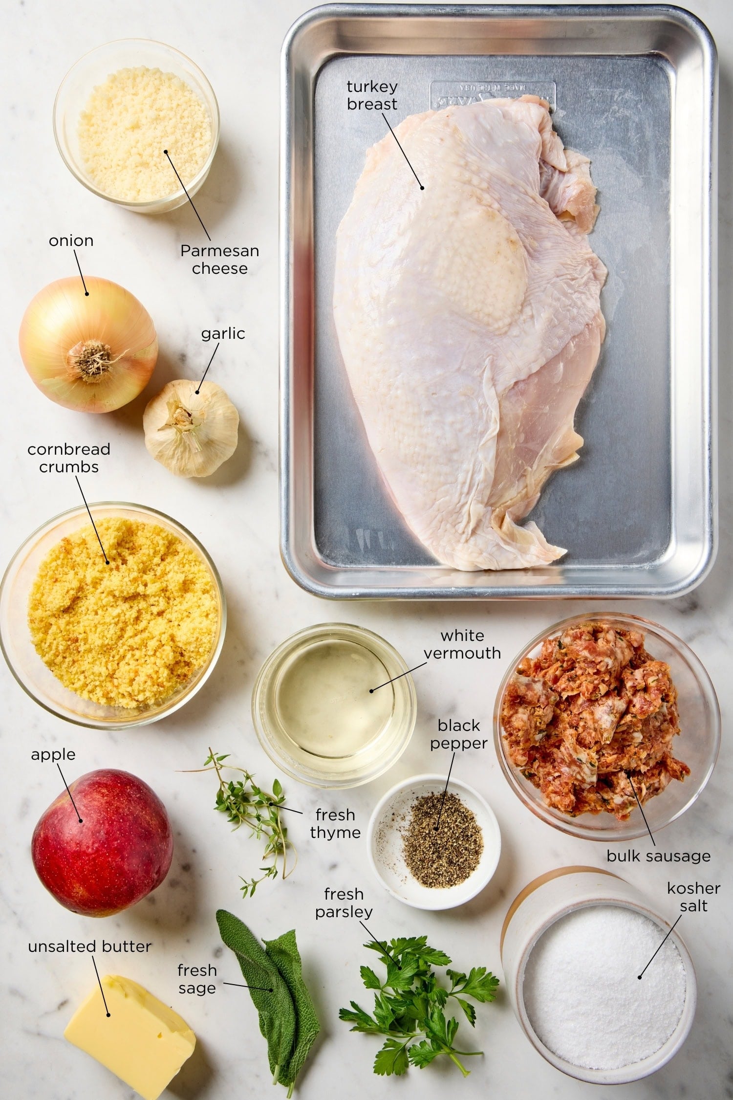 White surface filled with ingredients for turkey roulade including apple, sage, turkey breast, cornbread crumbs, and more.