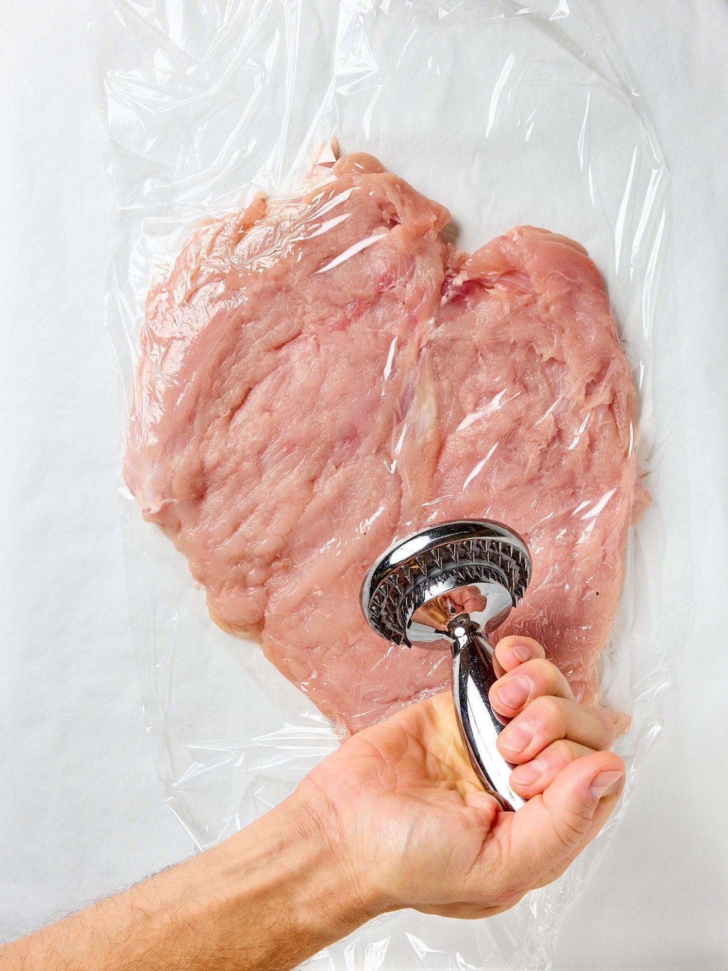 Hand pounding out a turkey breast on a piece of plastic wrap.