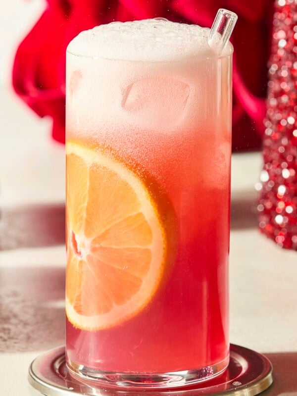 Tall glass with an orange slice down the side and pink color mocktail liquid.