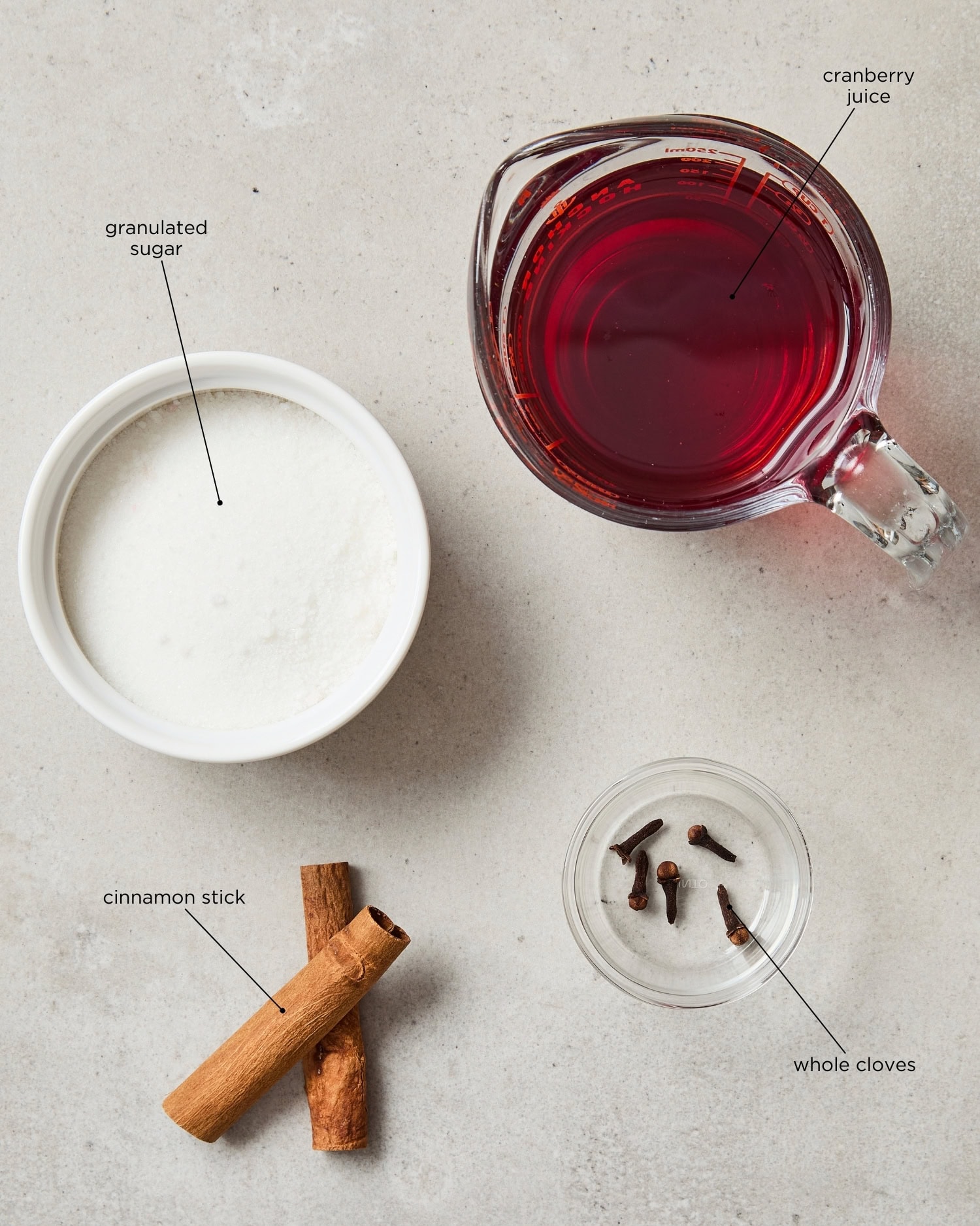 Concrete surface with ingredients needed for cranberry simple syrup including juice, sugar, cinnamon, and cloves.
