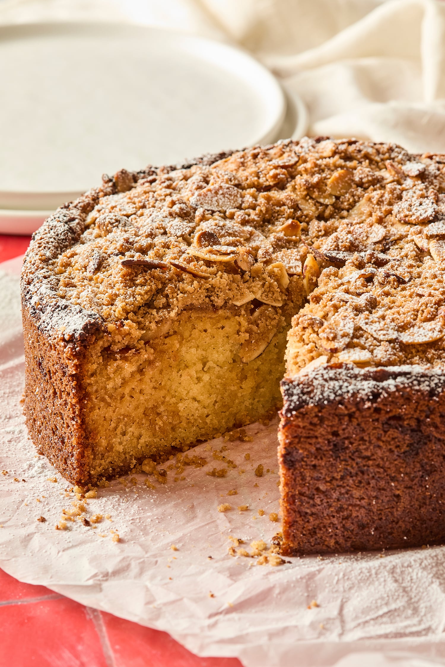 Slice taken out of a frangipane coffee cake with almond streusel topping with plates in background.
