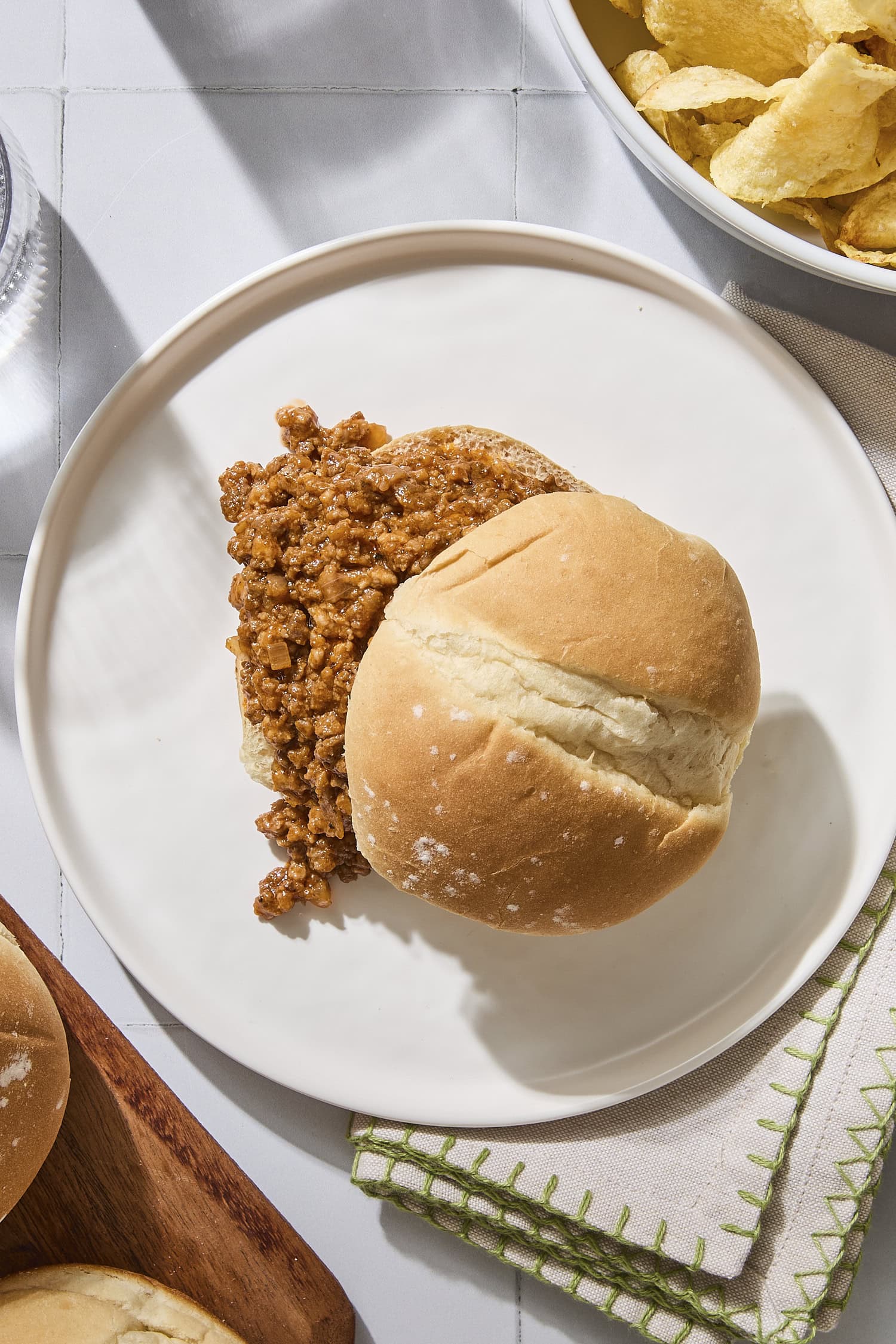 A hamburger bun with sloppy joe meat rests on a plate next to other buns, meat, and chips.