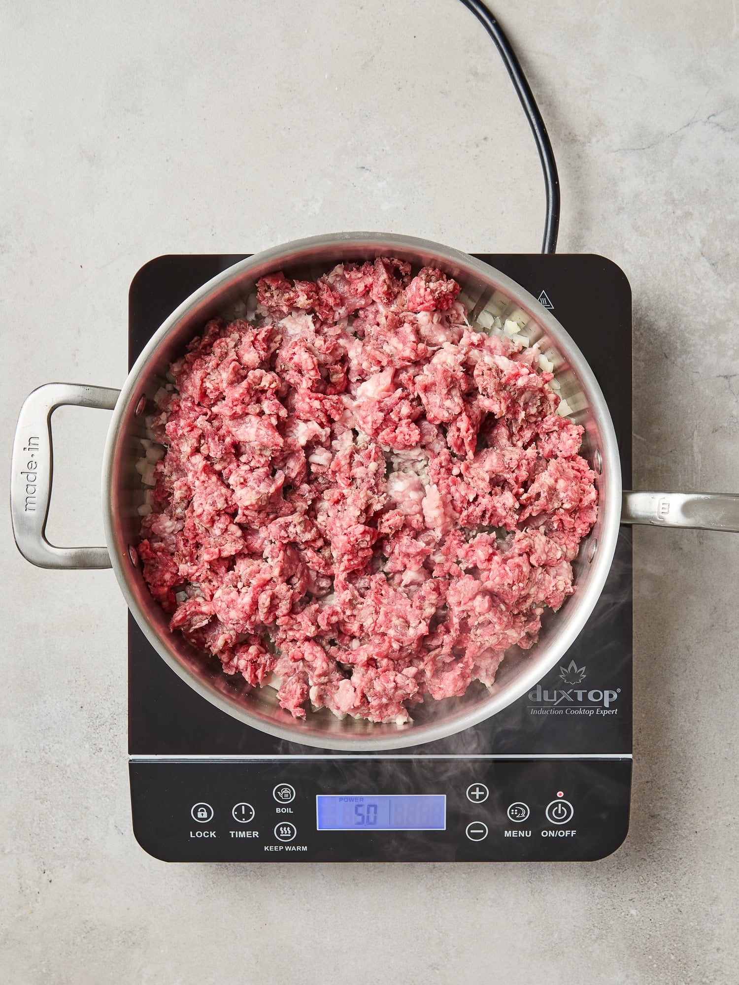 Ground pork and beef begin to cook in a pan on a stovetop.