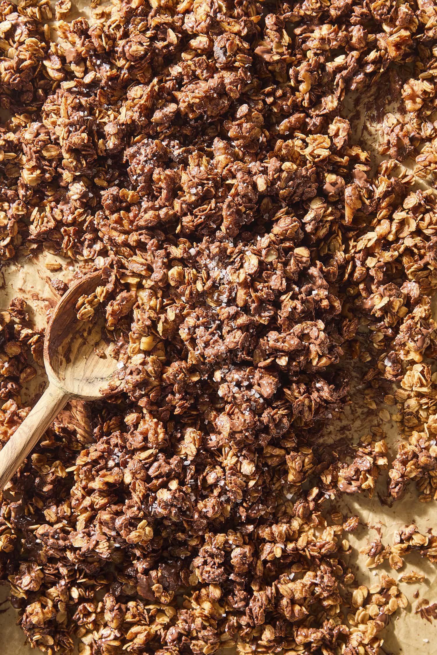 Spoon mixing pieces of chocolate chip cookie granola on baking sheet.