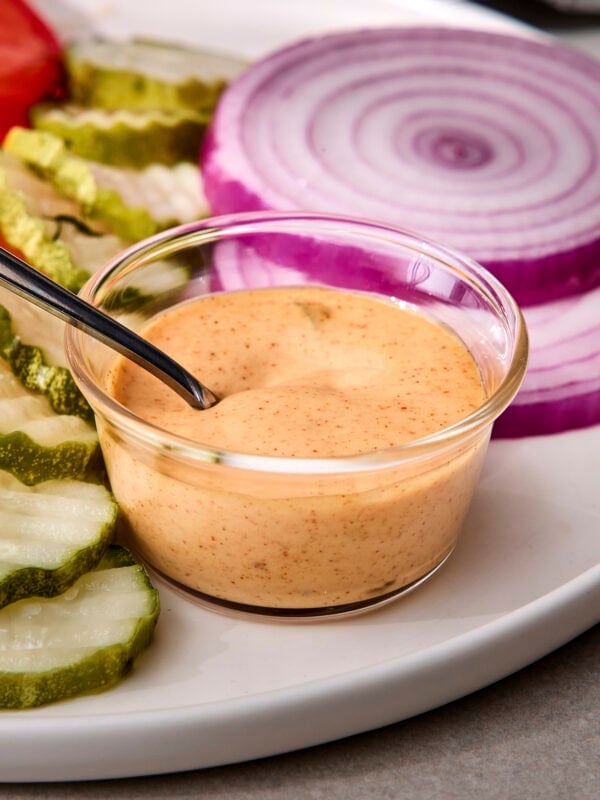 Homemade burger sauce in small glass bowl with pickles and red onion in background.