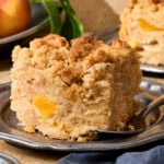 Slice of peach coffee cake on plate with peaches in background.