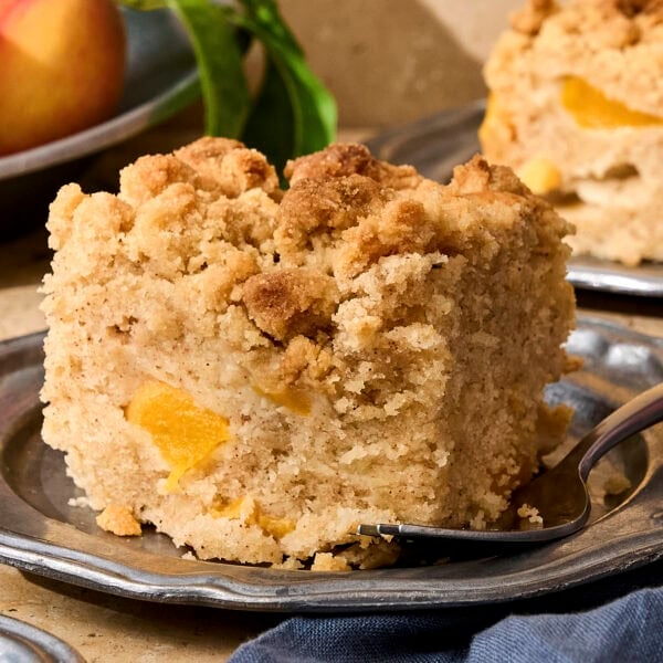 Slice of peach coffee cake on plate with peaches in background.