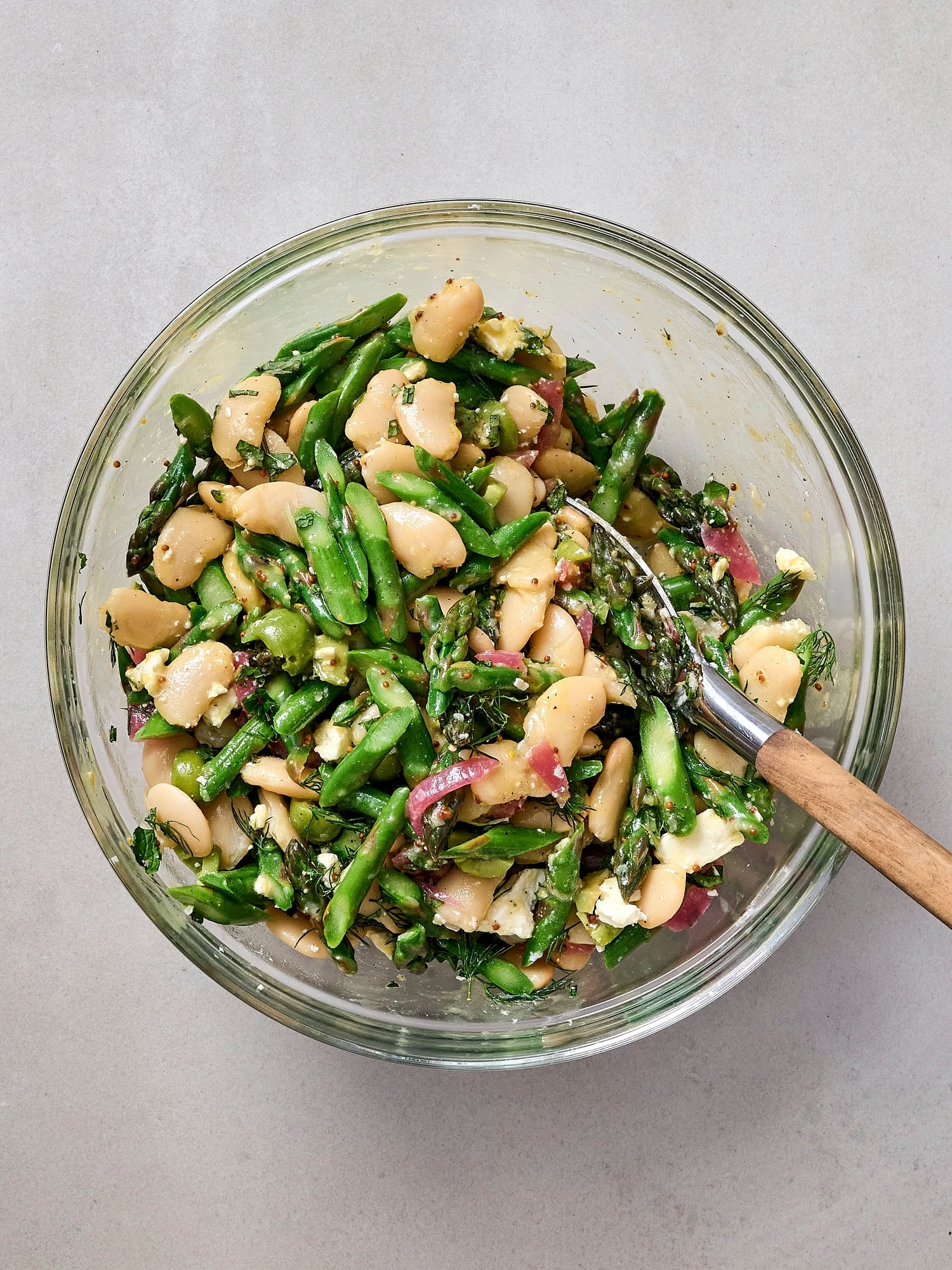 Glass bowl mixing together asparagus salad with white beans, feta, dill, tuna, and olives.
